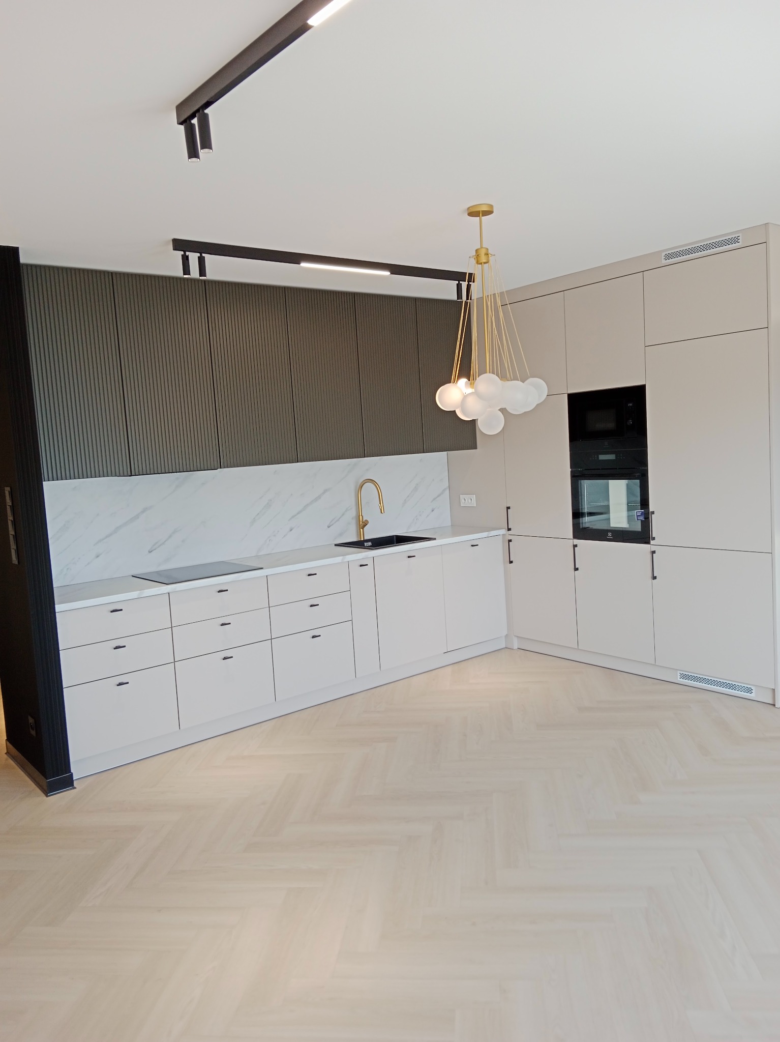 Nowoczesna kuchnia z jasnymi szafkami, złotą armaturą i marmurowym backsplashiem, oświetlona designerską lampą z białymi kulami, z widocznym parkietem ułożonym w jodełkę.