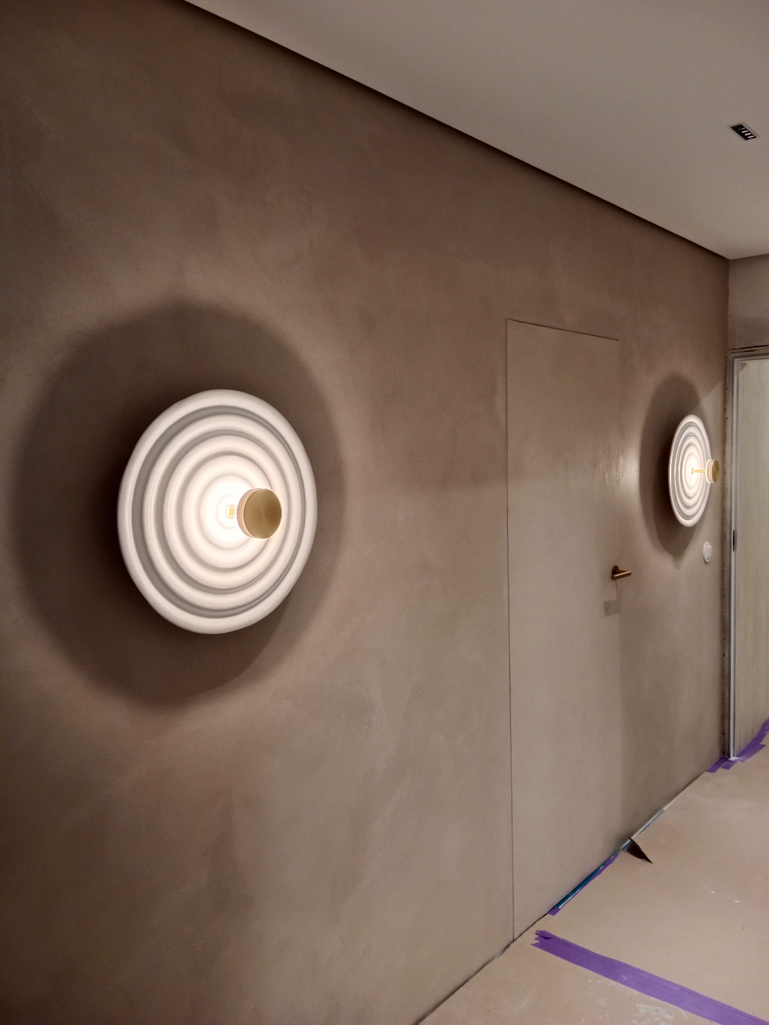 Ściana pokryta beżową, strukturalną farbą z dwoma okrągłymi lampami ściennymi z białymi koncentrycznymi okręgami, widoczne zabezpieczenia podłogi taśmą malarską.
