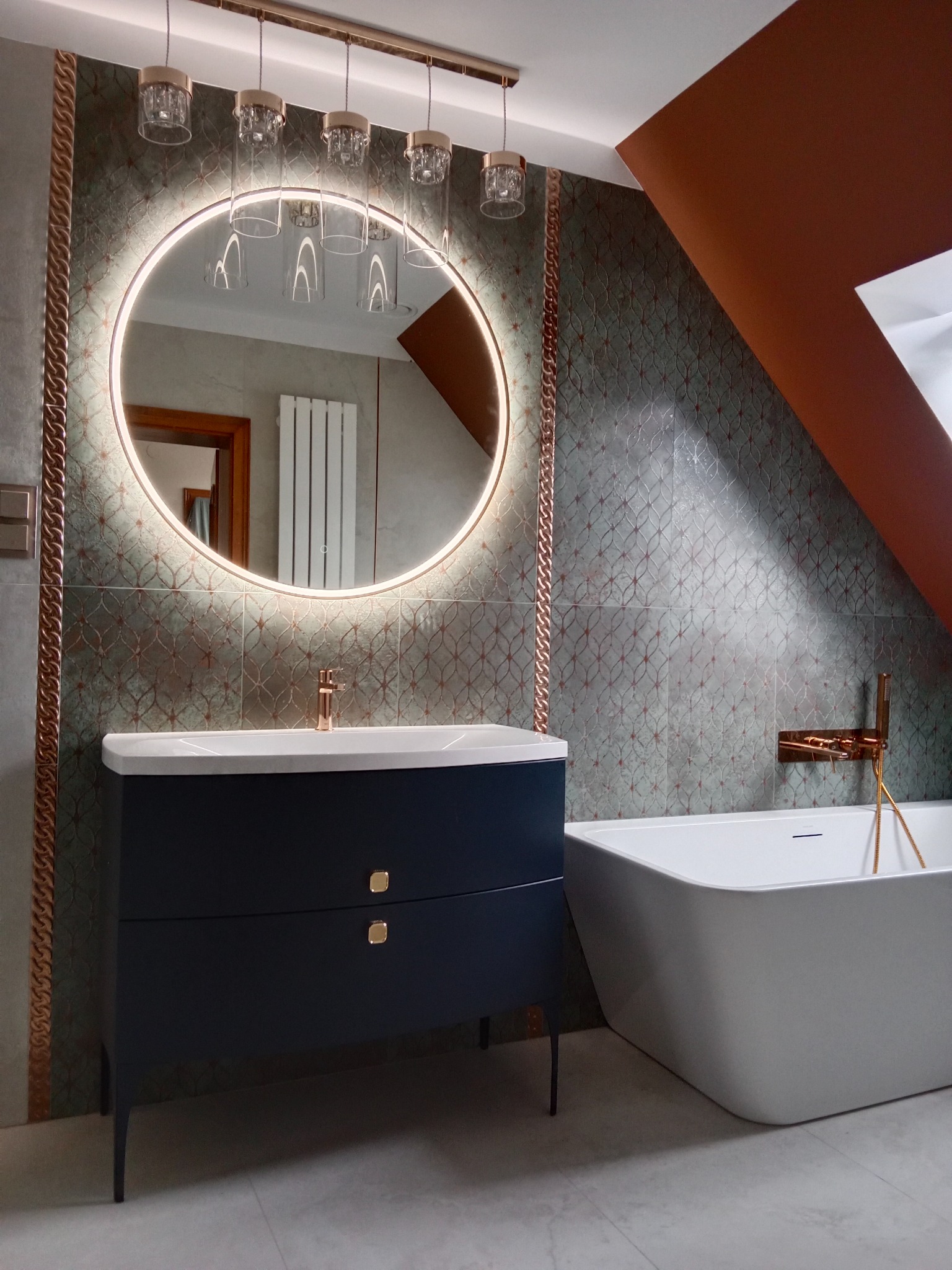 Nowoczesna łazienka z okrągłym lustrem LED, szafką pod umywalką w kolorze granatowym i białą wanną wolnostojącą. Uwagę przyciągają miedziane akcenty.