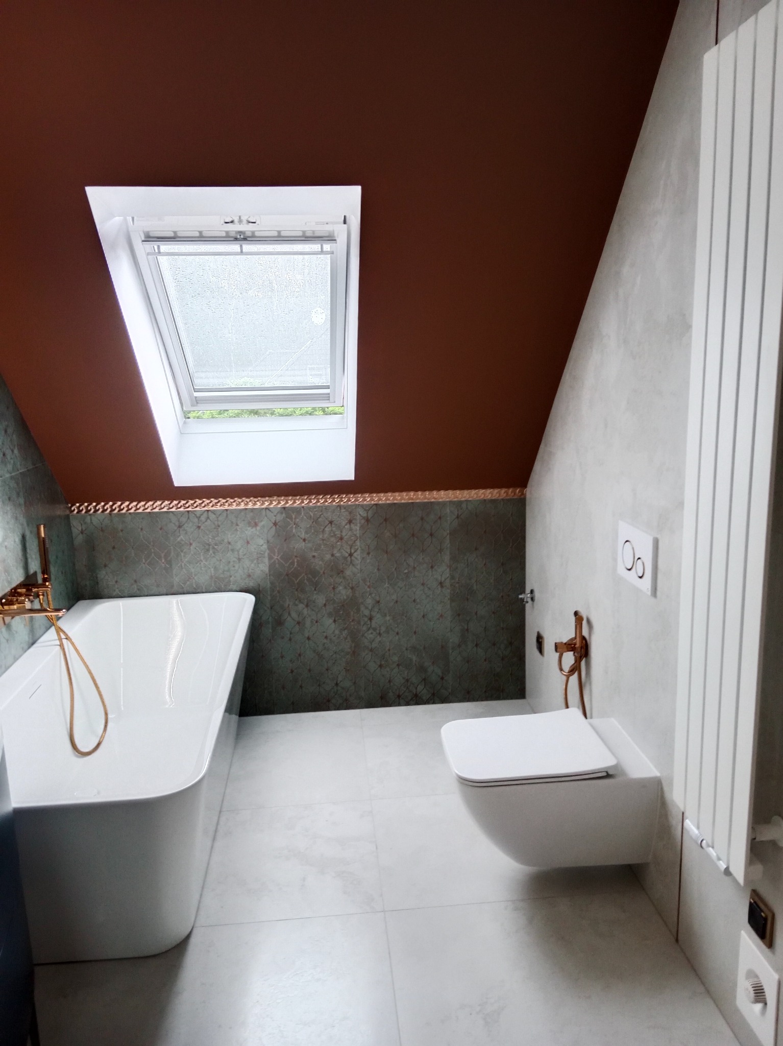 Stylowa łazienka na poddaszu z oknem dachowym, białą wanną i wiszącą toaletą. Ciemnobrązowy sufit kontrastuje z szarą ścianą i płytkami. Złote detale armatury dodają elegancji.