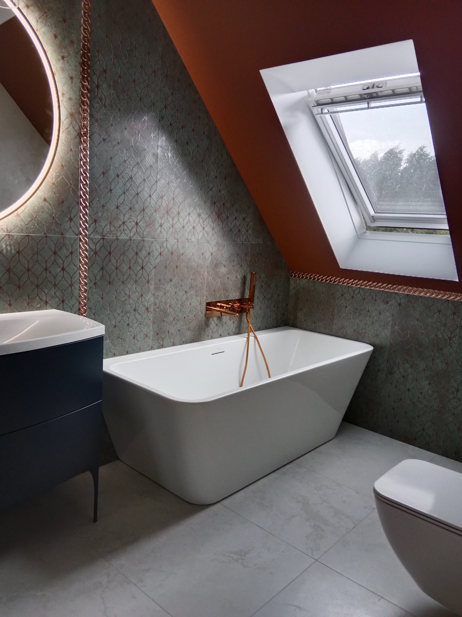 Nowoczesna łazienka na poddaszu z białą wanną wolnostojącą, umywalką na szafce i toaletą. Uwagę przykuwają zielono-miedziane płytki i okno dachowe.