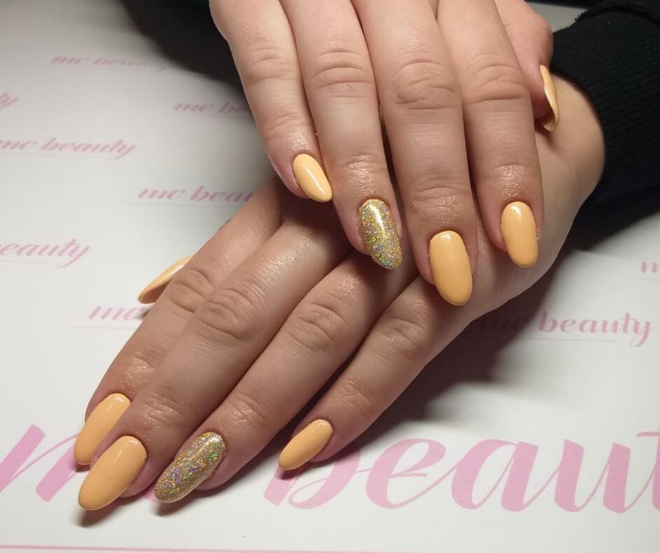 Dłonie z pomarańczowymi paznokciami hybrydowymi, jeden paznokieć na każdej dłoni pokryty brokatem na tle blatu z logo salonu kosmetycznego.