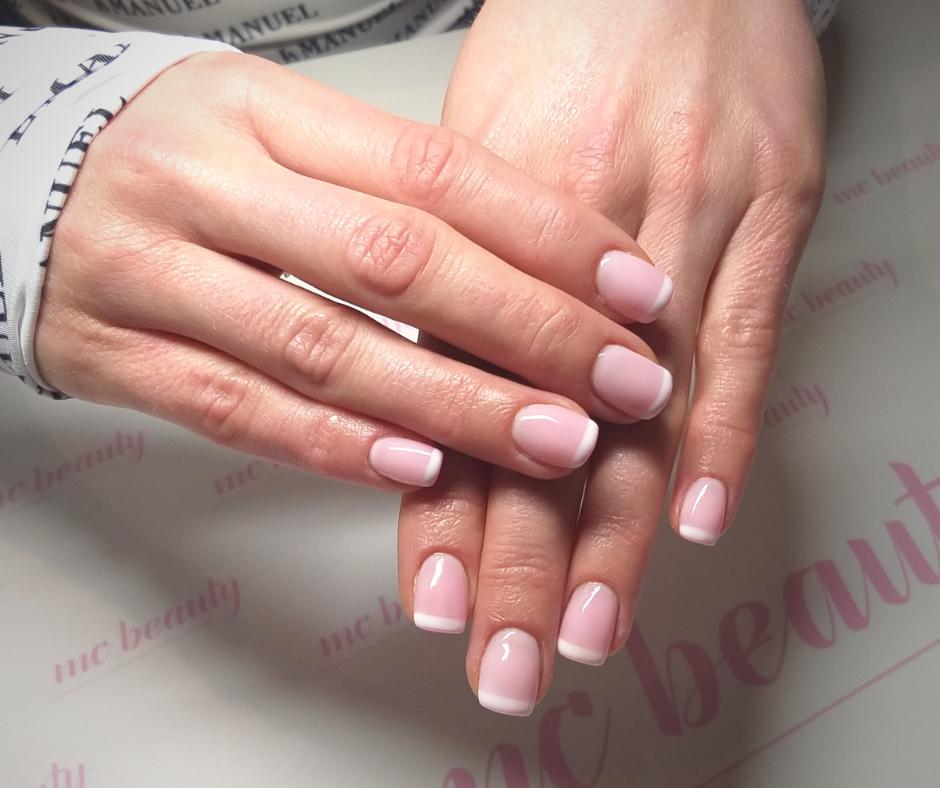 Dłonie z delikatnym, różowym manicure francuskim na tle blatu z subtelnym logo salonu kosmetycznego.