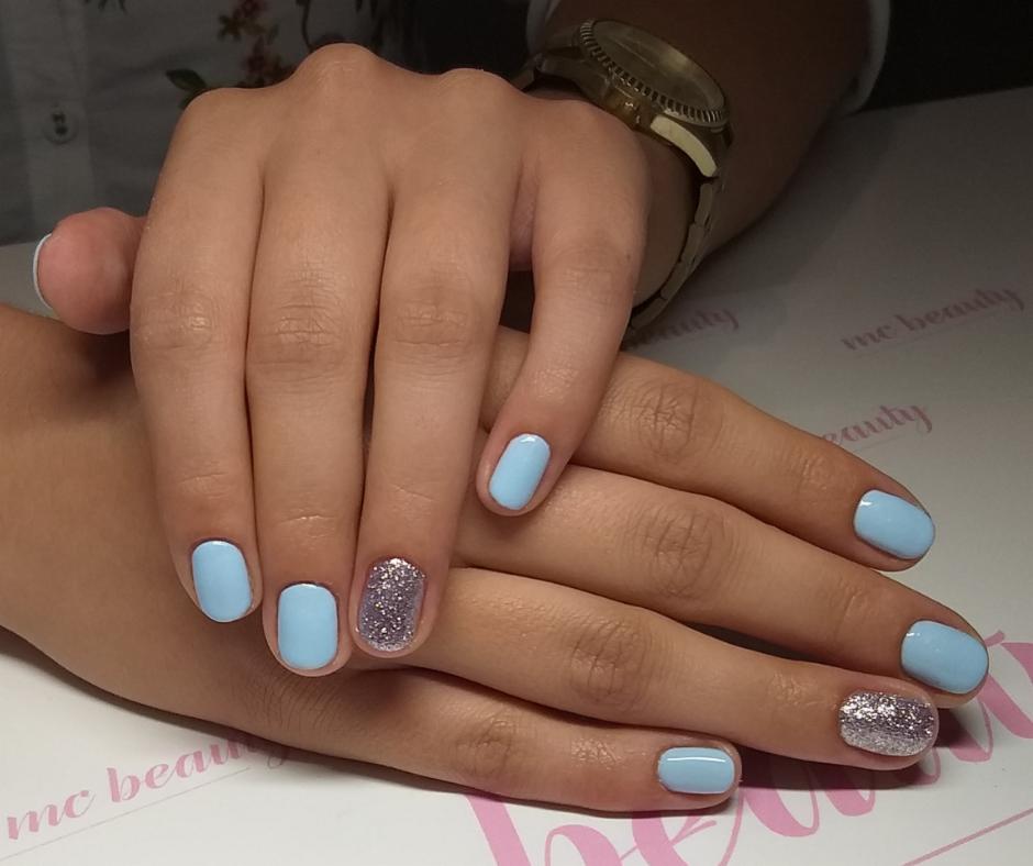Dłonie z jasnoniebieskim manicure hybrydowym, jeden paznokieć na każdej dłoni pokryty srebrnym brokatem, złoty zegarek na nadgarstku.