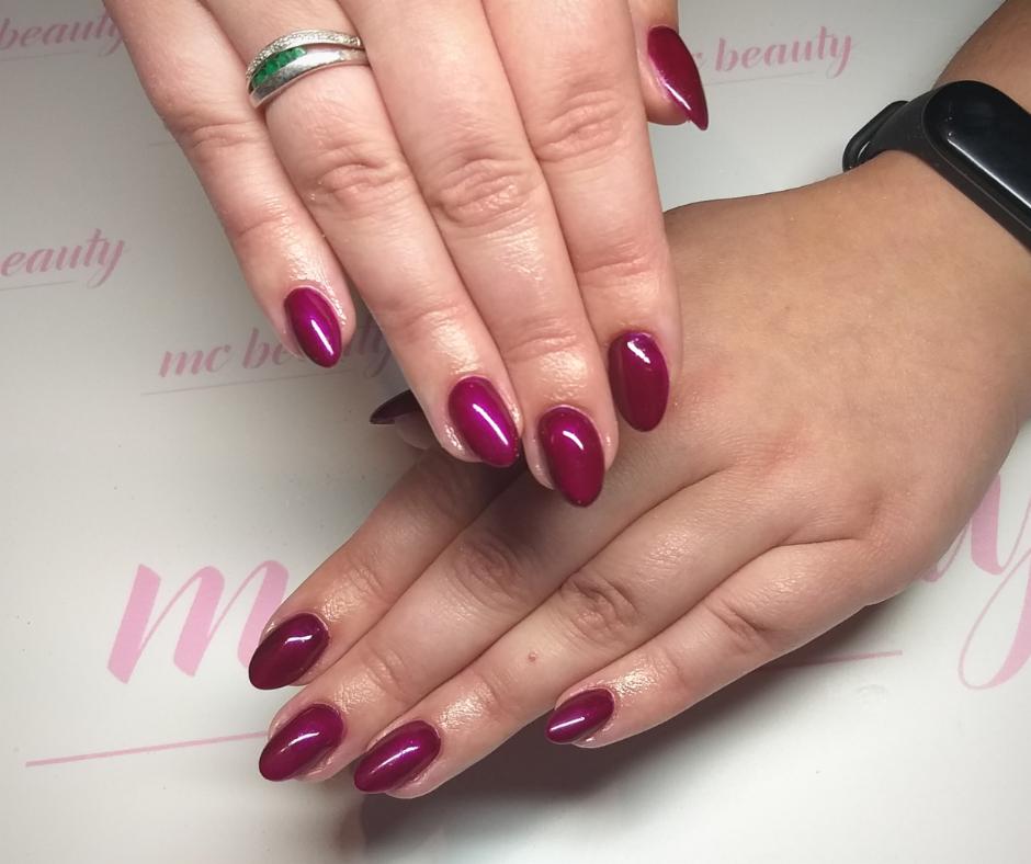 Dłonie z bordowymi, błyszczącymi paznokciami w kształcie migdała, na jednej dłoni pierścionek z zielonym oczkiem, tło z delikatnym różowym napisem.