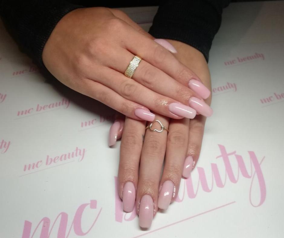 Dłonie z jasnoróżowym, błyszczącym manicure hybrydowym o kształcie migdała, zdobione pierścionkami: szerokim złotym z cyrkoniami i delikatnym srebrnym w kształcie serca, na tle z logo salonu...