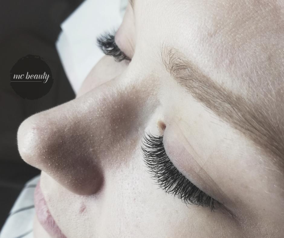 Zbliżenie na oko z przedłużonymi, gęstymi rzęsami; widoczny pieprzyk na skroni i logo 'mc beauty' w lewym górnym rogu.