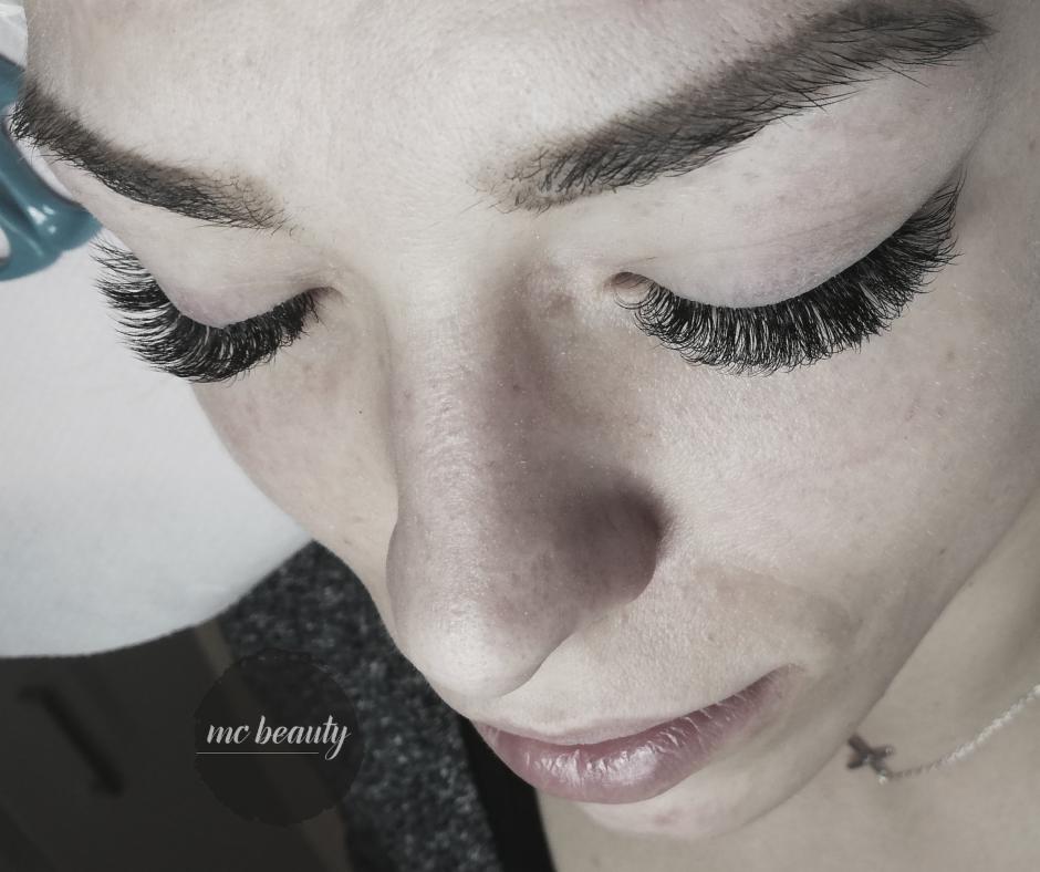 Zbliżenie na twarz kobiety z wyraźnie podkreślonymi, długimi rzęsami po zabiegu przedłużania. Widoczna delikatna biżuteria na szyi i logo firmy w lewym dolnym rogu.