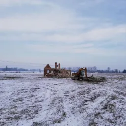 Pomarańczowa koparka na zasypanym śniegiem polu, w tle ruiny ceglanego budynku z kominami i zwały gruzu.