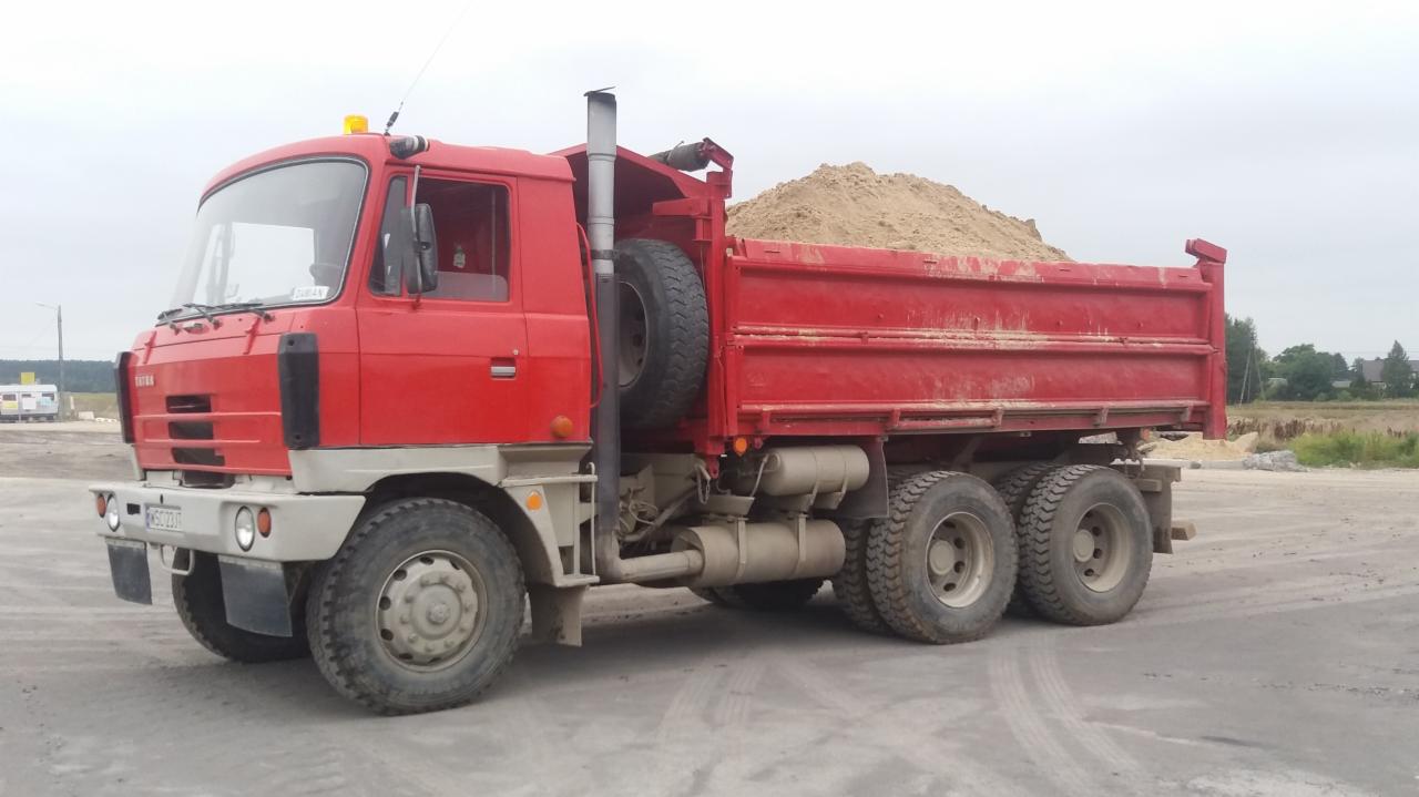 Czerwona wywrotka Tatra z ładunkiem piasku na placu budowy, widoczne ślady opon na szarym podłożu.