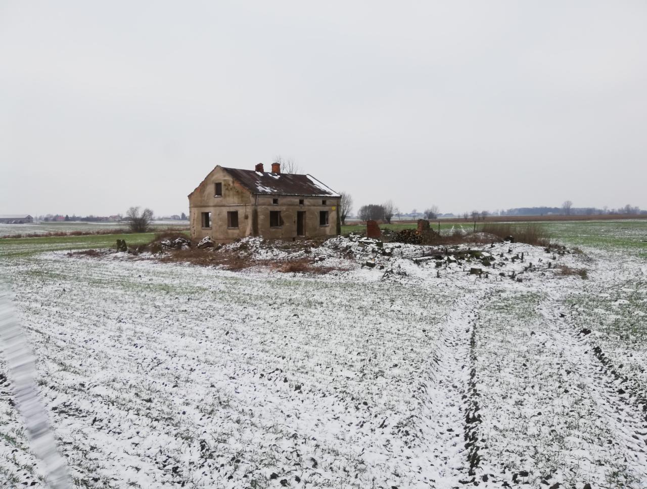 Zrujnowany, częściowo rozebrany budynek w zimowym krajobrazie, otoczony śniegiem i gruzem, z widocznymi śladami opon na pierwszym planie.