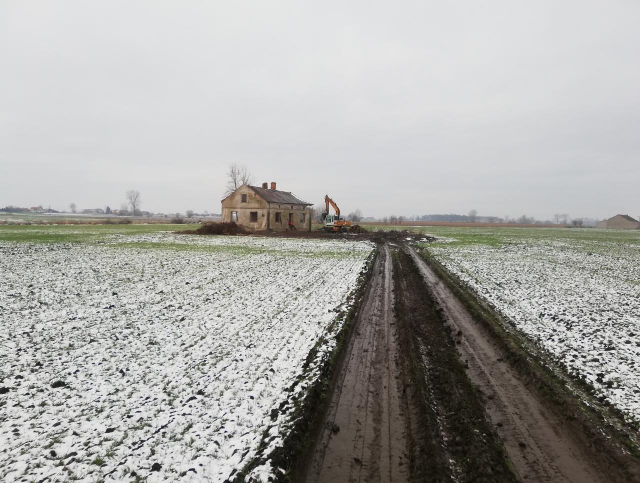 Krajobraz polny z resztkami śniegu, zniszczony budynek w oddali i pomarańczowa koparka wykonująca prace ziemne, widoczne ślady opon w błocie.