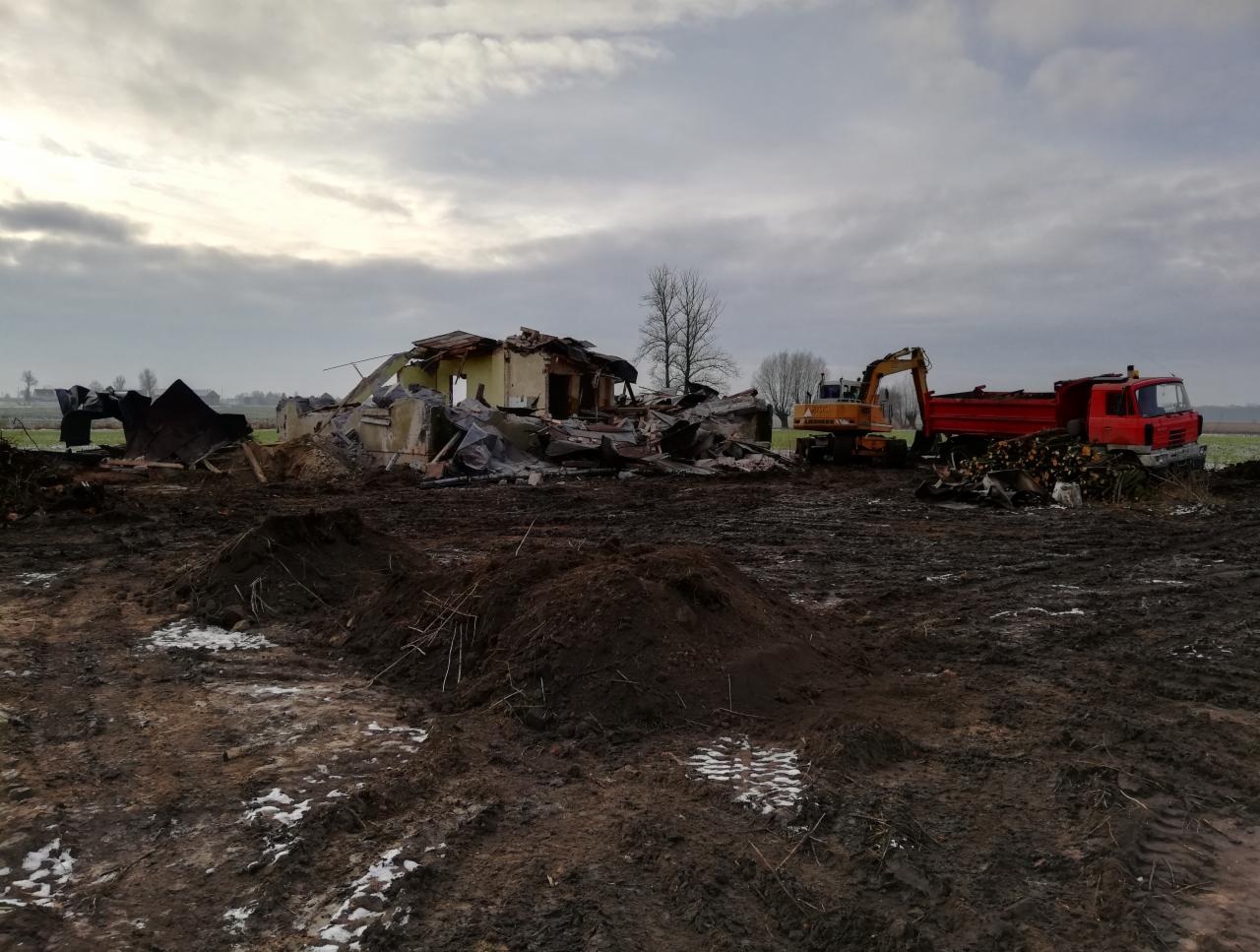 Rozbiórka budynku w toku, z widocznym gruzem, koparką i ciężarówką na błotnistym terenie w pochmurny dzień.