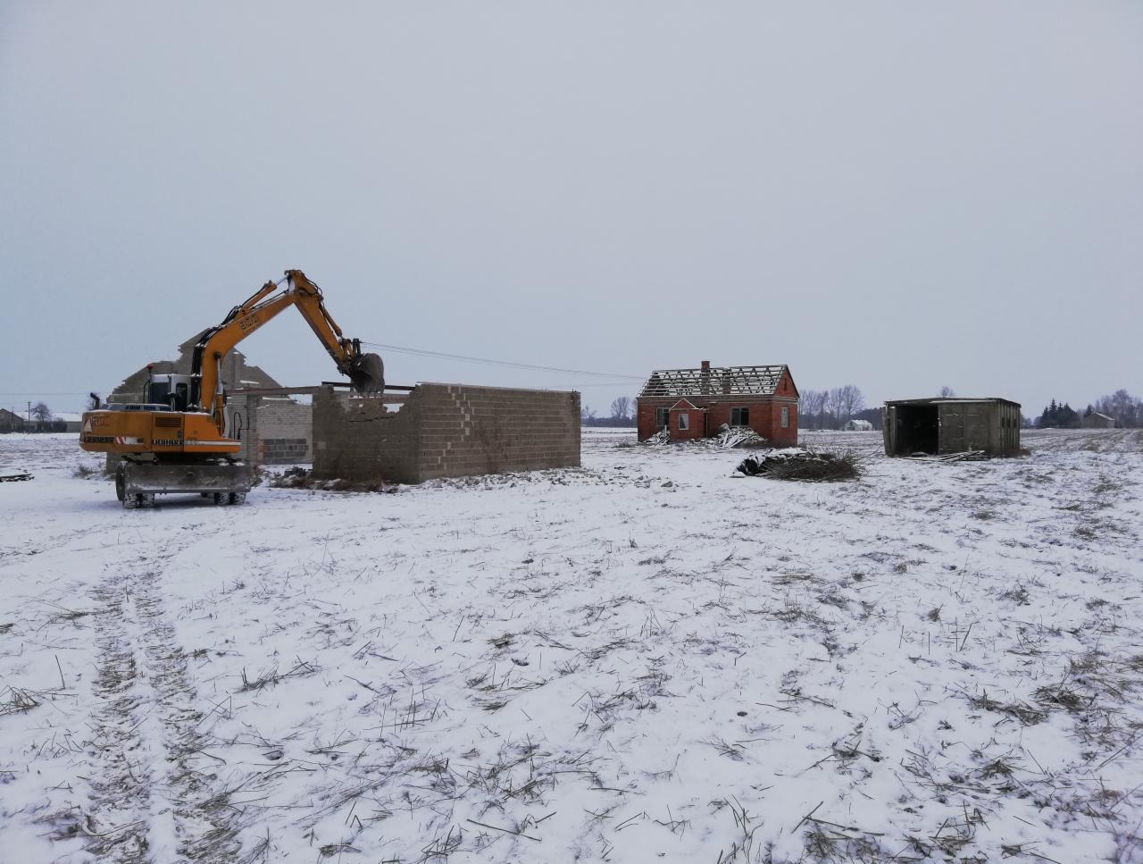 Żółta koparka Liebherr 500 rozbiera resztki murowanej konstrukcji na zasypanym śniegiem polu, w tle zrujnowany, ceglany budynek bez dachu.