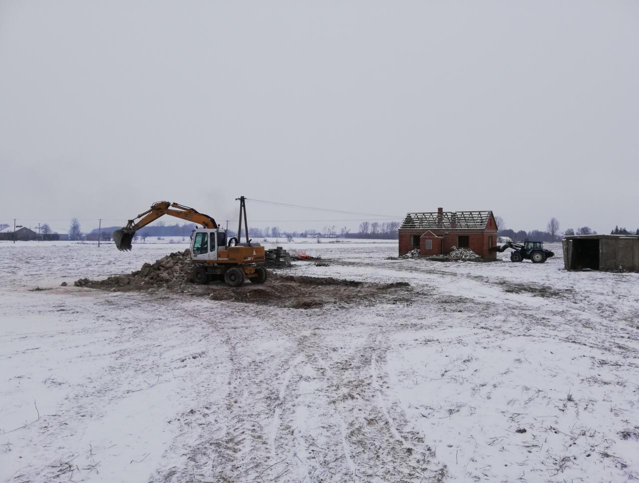 Żółta koparka nabiera ziemię w wykopie na zasnieżonym polu, w tle niedokończony budynek z czerwonej cegły i traktor.