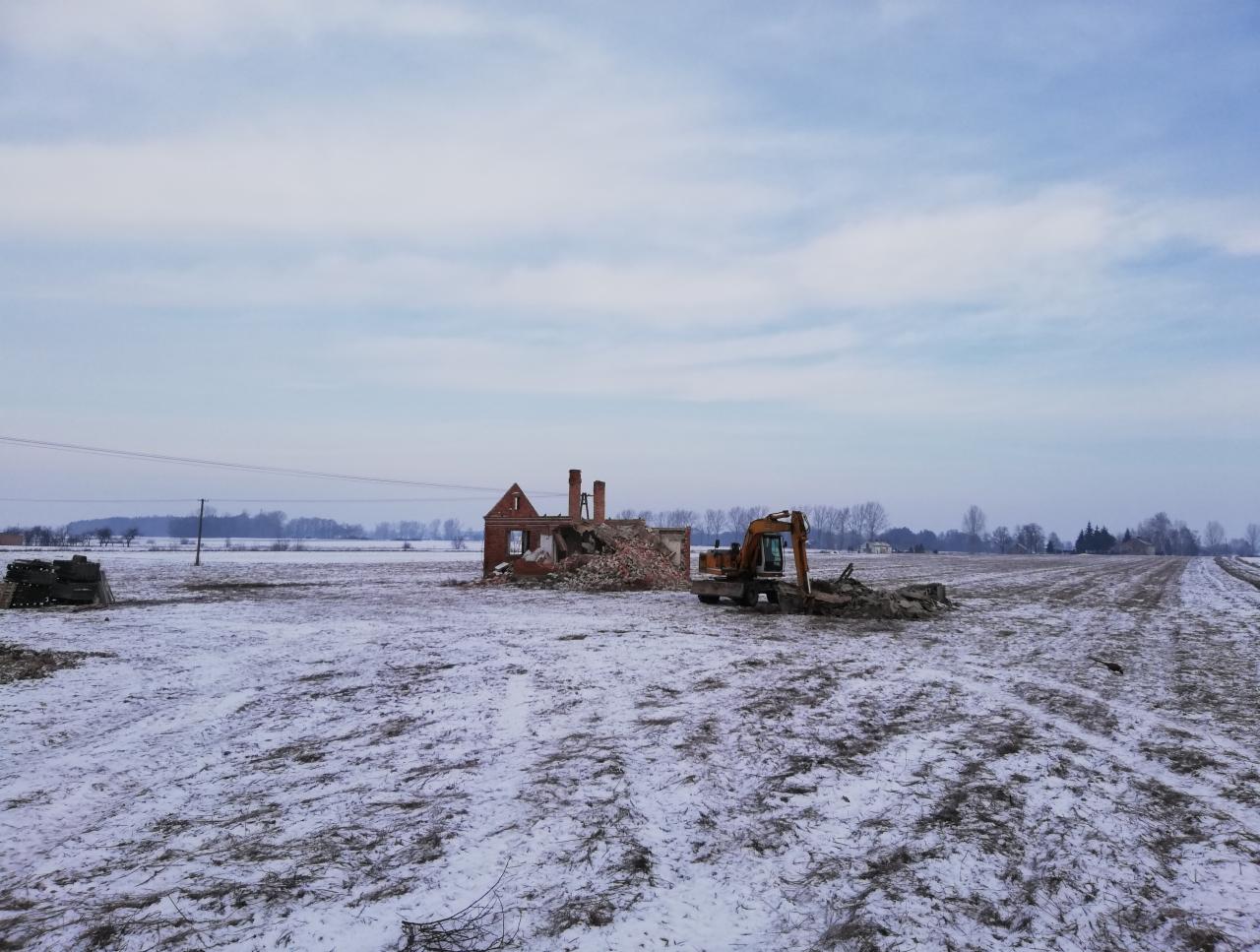 Pomarańczowa koparka na zasypanym śniegiem polu, w tle ruiny ceglanego budynku z kominami i zwały gruzu.