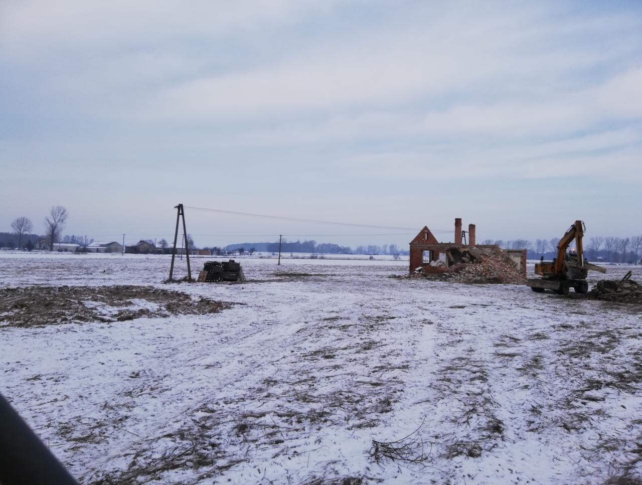 Pomarańczowy koparka przy częściowo zburzonym ceglanym budynku na zaśnieżonym polu, słupy energetyczne w tle.