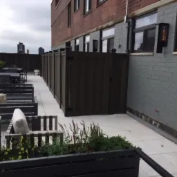 Nowoczesny taras na dachu z drewnianą zabudową, białymi płytami i meblami ogrodowymi w odcieniach szarości oraz roślinami w donicach.