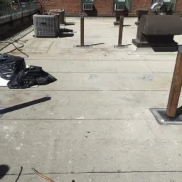 Płaski dach z widocznymi elementami instalacji, rurami, wentylacją i workami na śmieci, zarys budynku w tle.