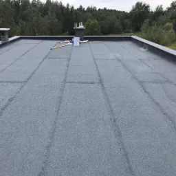 Płaski dach pokryty papą termozgrzewalną z widocznymi łączeniami, kominy wentylacyjne i narzędzia dekarskie na powierzchni, w tle drzewa.