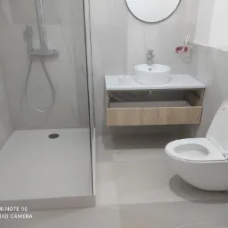 Nowoczesna łazienka z kabiną prysznicową, wiszącą toaletą i umywalką nablatową na szafce z otwartą szufladą, wykończona jasnymi płytkami imitującymi marmur.