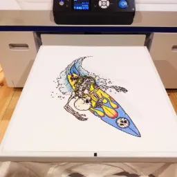 Koszulka z nadrukiem szkieletu surfera na desce, wychodząca z drukarki do tekstyliów, widok z góry, oświetlenie studyjne.