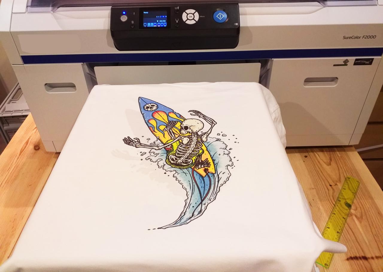 Biała koszulka z nadrukiem szkieletu na desce surfingowej, umieszczona pod drukarką SureColor F2000, widoczny fragment drewnianego blatu i żółta miarka.