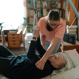 Medicus Katarzyna Dembs - Kobieta w okularach pomaga starszemu mężczyźnie leżącemu w łóżku, używając podnośnika. W tle regał z książkami i inne meble w pokoju.
