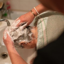 Medicus Katarzyna Dembs - Mycie głowy seniora w zlewie przez opiekuna. Widoczne spienione włosy, ręce z biżuterią i fragment ręcznika. Domowa łazienka z kranem w tle.