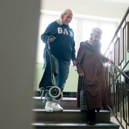 Medicus Katarzyna Dembs - Opiekunka z wózkiem zakupowym i starsza kobieta schodzą po schodach. Klatka schodowa z metalową barierką. Światło dzienne z okna w tle.