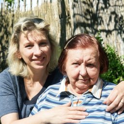 Medicus Katarzyna Dembs - Uśmiechnięta kobieta obejmuje starszą panią na tle zieleni i drewnianego płotu. Przyjazna atmosfera, opieka i wsparcie w słoneczny dzień.