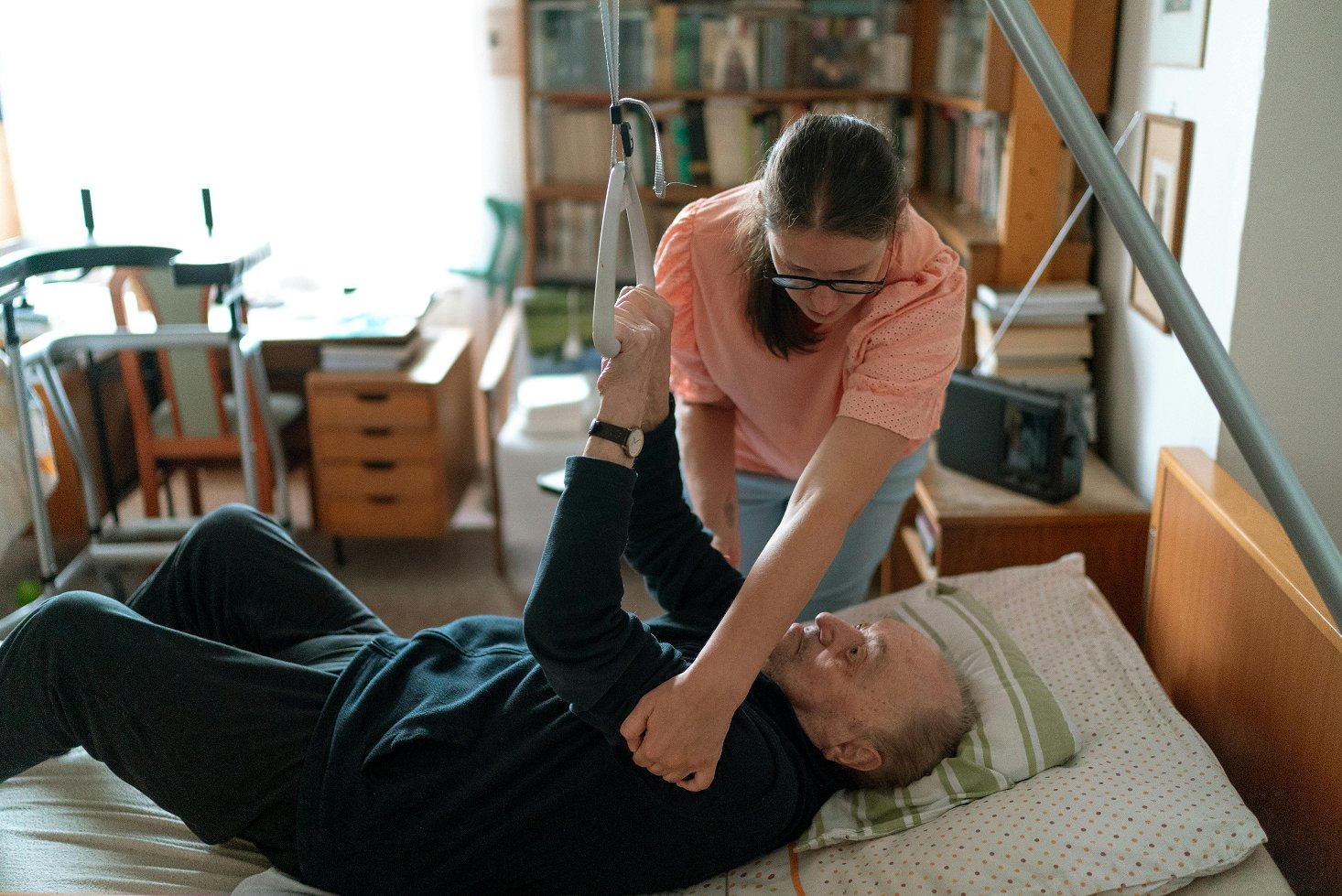 Kobieta w okularach pomaga starszemu mężczyźnie leżącemu w łóżku, używając podnośnika. W tle regał z książkami i inne meble w pokoju.