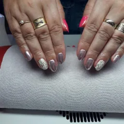 Dłonie z różnymi stylizacjami paznokci: metaliczne, różowe szpiczaste i z motywem róży, zdobione pierścionkami na tle białej podkładki.