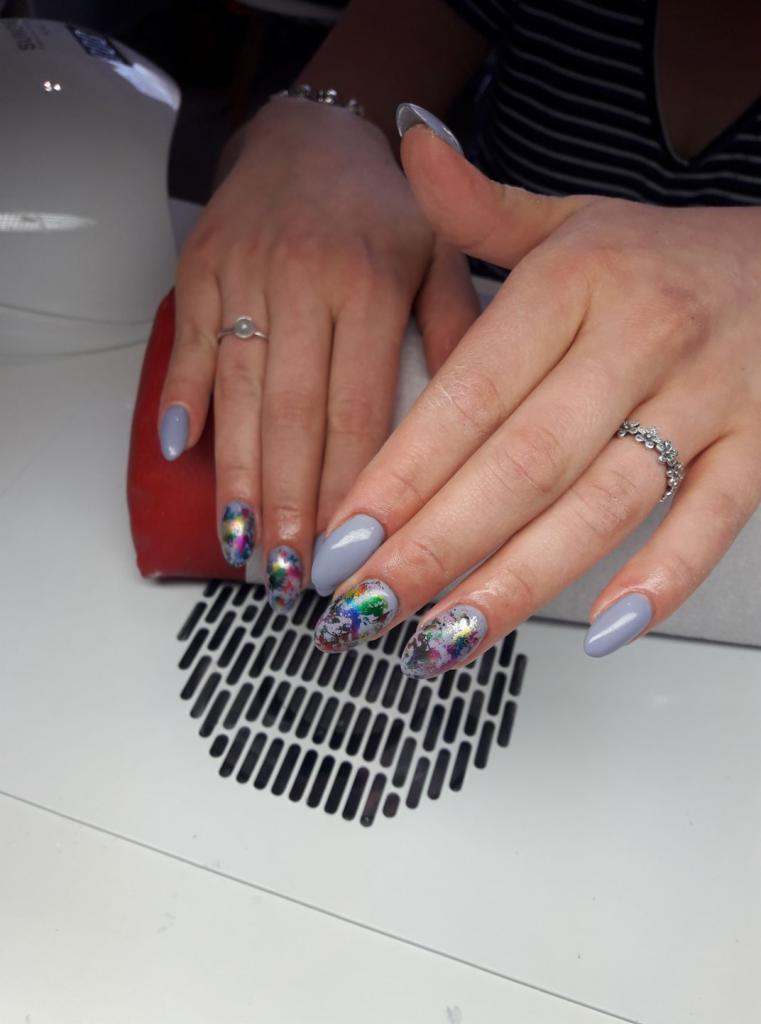 Dłonie z szarymi paznokciami w kształcie migdała, dwa z nich ozdobione folią z efektem benzyny, widoczne pierścionki i element pochłaniacza pyłu.