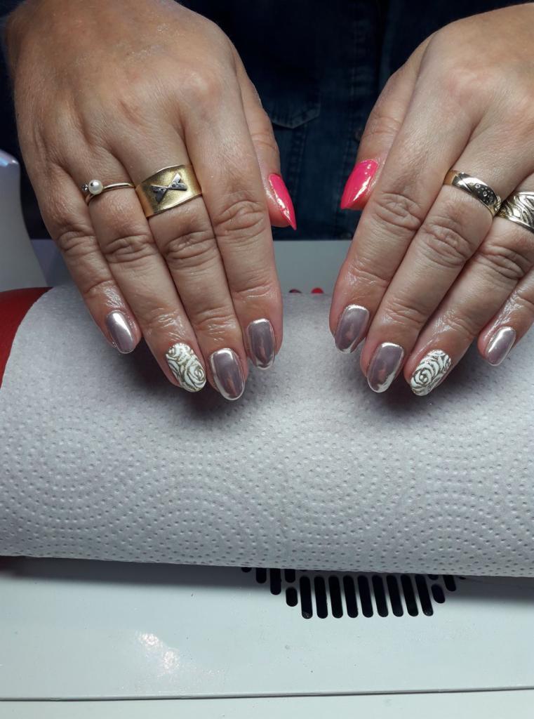 Dłonie z różnymi stylizacjami paznokci: metaliczne, różowe szpiczaste i z motywem róży, zdobione pierścionkami na tle białej podkładki.