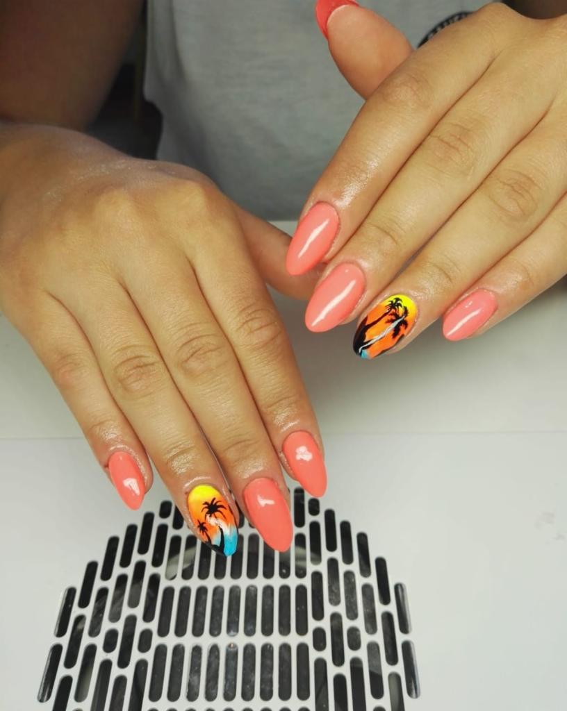 Dłonie z pomarańczowymi, migdałowymi paznokciami; na dwóch paznokciach widnieje motyw zachodu słońca z palmami.