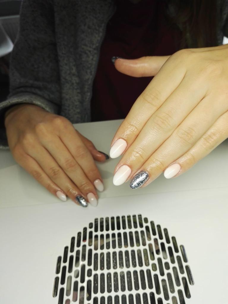 Dłonie z świeżo pomalowanymi paznokciami w odcieniach bieli i srebra, manicure hybrydowy z efektem brokatu na jednym paznokciu, na tle lampy UV do paznokci.