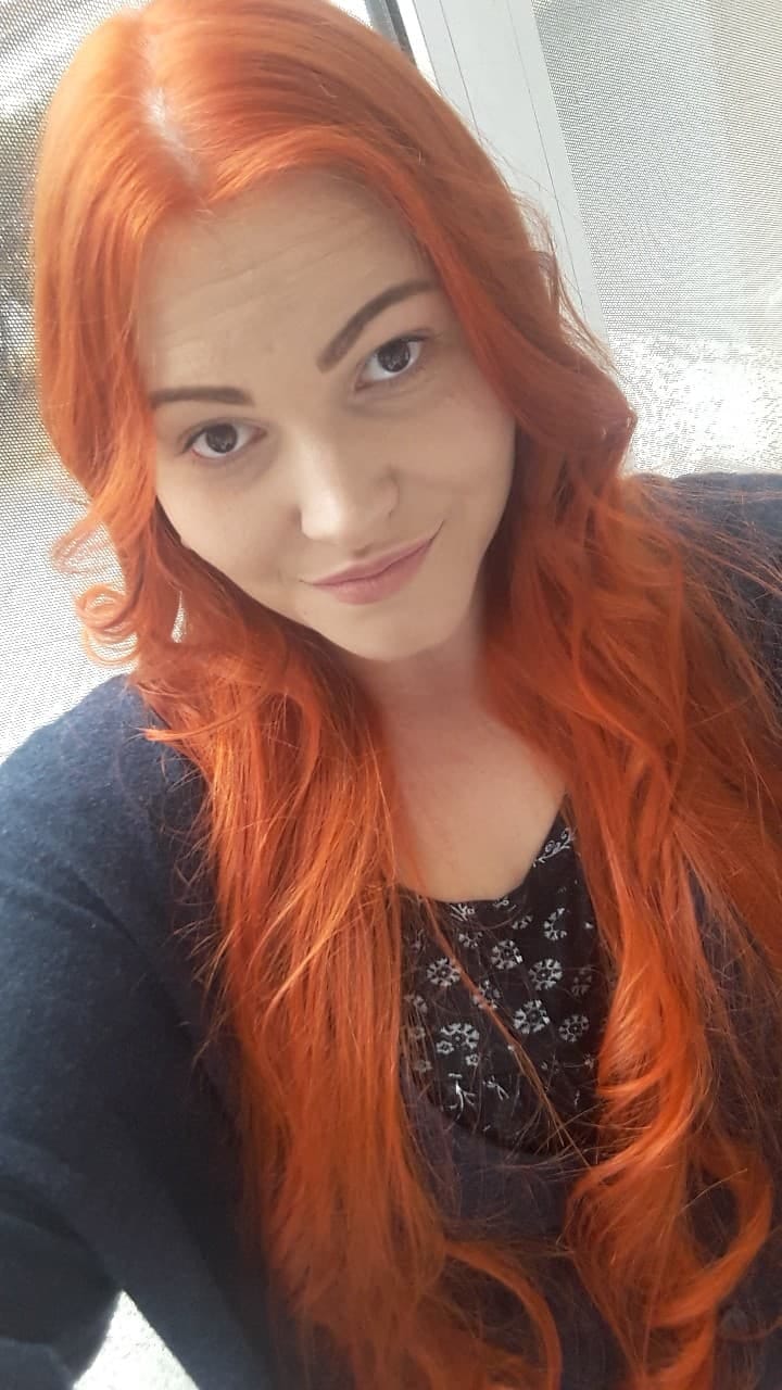 Portret kobiety z długimi, rudymi włosami, delikatny uśmiech, ciemne oczy, ujęcie z bliska, widoczne pojedyncze kosmyki włosów.