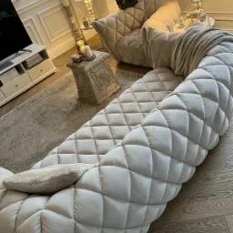 Luksusowy salon z beżową, modułową sofą w romby, na kt&oacute;rej leży poduszka, obok stolik z dekoracjami i dywan, w tle fotel z szarym kotem i lampy.