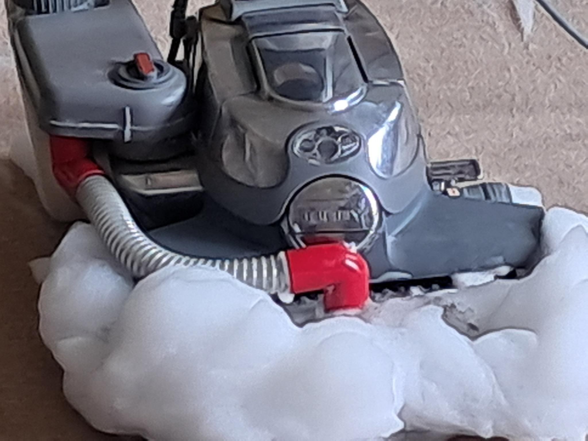 Szary odkurzacz piorący Kirby w trakcie pracy, otoczony pianą czyszczącą na beżowym tle. Widoczny czerwony element łączący rurę.