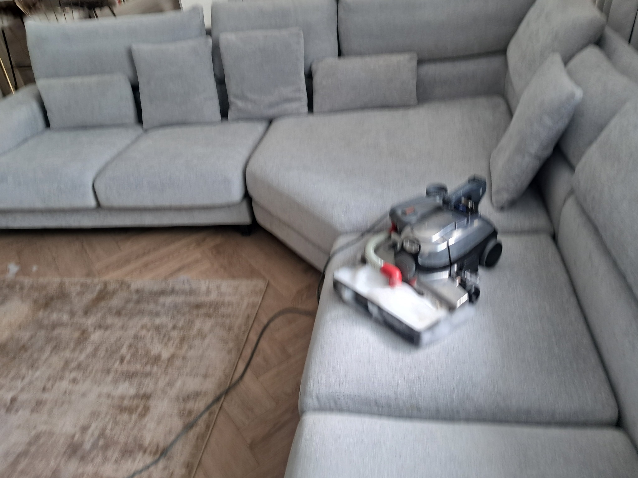 Jasny, narożny, modułowy narożnik po praniu. Na siedzisku stoi odkurzacz piorący. Widoczne ślady po praniu na jasnej tapicerce. W tle poduszki i dywan.