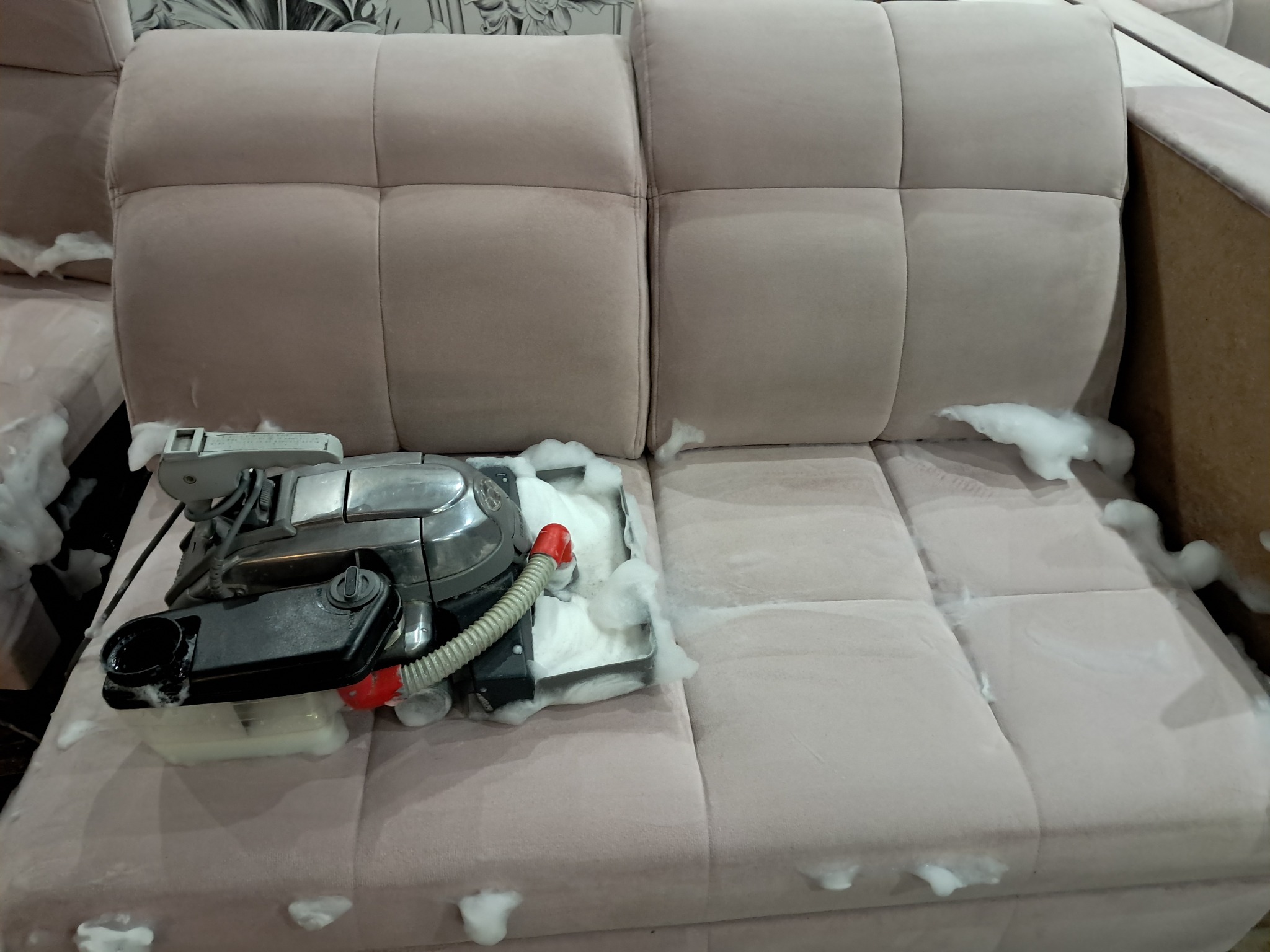 Beżowa kanapa z widocznym fragmentem czyszczonej powierzchni i pianą. Na kanapie stoi odkurzacz piorący z widocznym zbiornikiem na wodę i wężykiem.