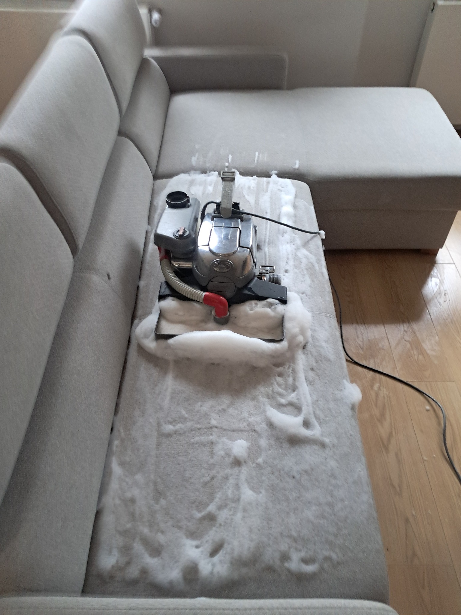 Szary narożnik czyszczony parowo-odkurzaczem z użyciem piany. Widoczny fragment podłogi. Sprzęt do czyszczenia tapicerki w trakcie pracy.