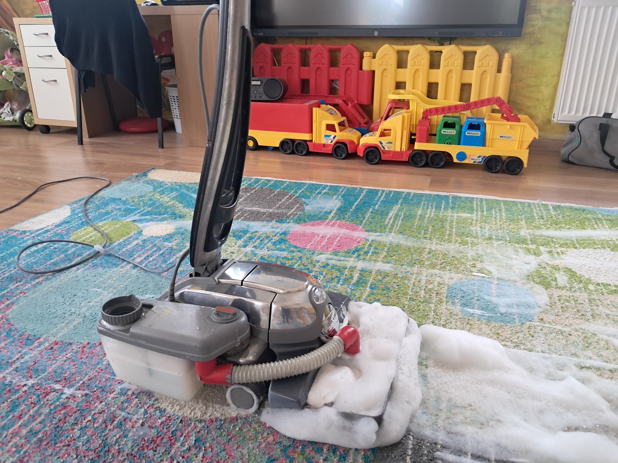 Pranie dywanu odkurzaczem piorącym z widoczną pianą. W tle zabawki dziecka i meble. Usługa prania dywanów w domu klienta.