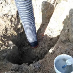 Czarna karbowana rura drenażowa w wykopie w ziemi, obok wiadro z zaprawą, Bartoszyce.