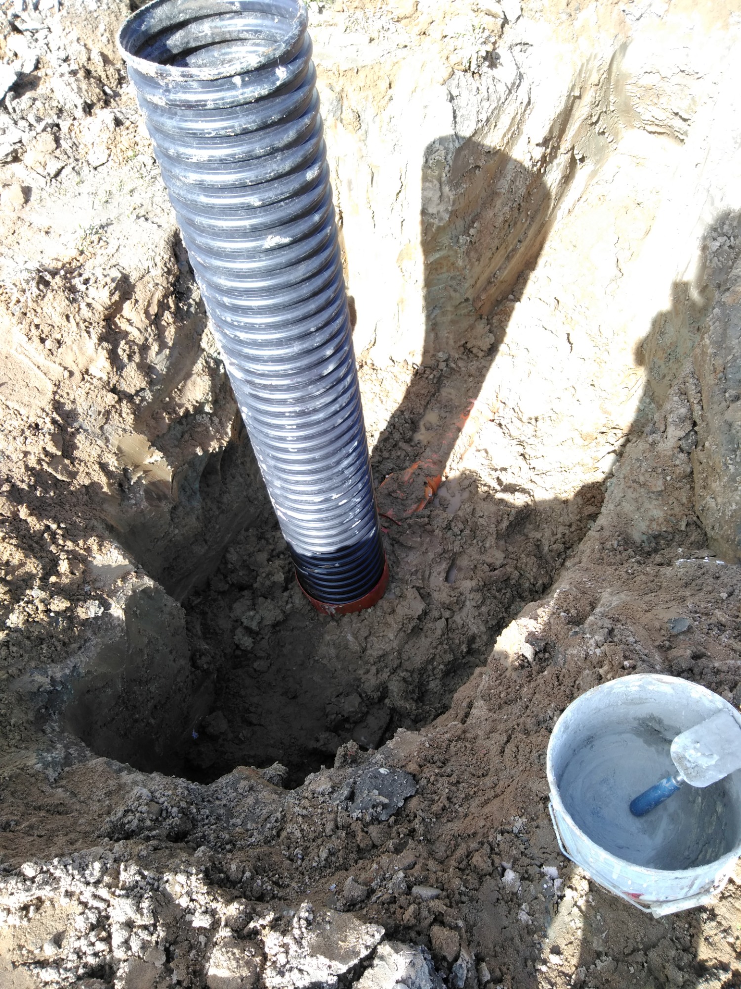 Czarna karbowana rura drenażowa w wykopie w ziemi, obok wiadro z zaprawą, Bartoszyce.