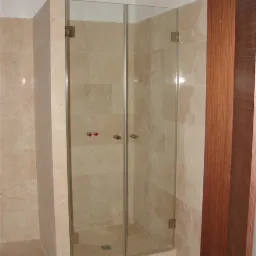 Szklana kabina prysznicowa na wymiar z dwuskrzydłowymi drzwiami, zamontowana w łazience wyłożonej beżowymi płytkami marmuropodobnymi, widok z zewnątrz kabiny.