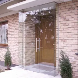 Szklana zabudowa wejścia do domu z cegły, widoczne drzwi z hartowanego szkła z długimi, pionowymi uchwytami.