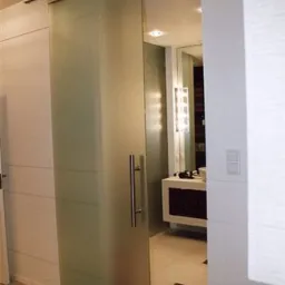 Matowe, szklane drzwi przesuwne z metalowym uchwytem i szyną górną, oddzielające łazienkę z widoczną umywalką i oświetleniem lustra.