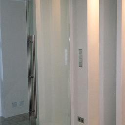 Drzwi Szklane Kabiny Prysznicowe ART-MAR - Szklane drzwi przesuwne z satynowym wykończeniem, zawieszone na metalowej szynie z widocznymi rolkami, z długim, pionowym uchwytem ze stali nierdzewnej, na tle białej ściany z włącznikami...